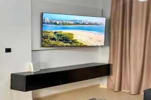 A flat-screen TV mounted on a wall above a black entertainment unit, with a beach scene on the screen. - Ground living (Tunis)