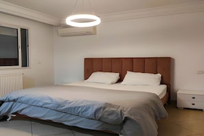 A bedroom with a bed, a bedside table, a nightstand, a window, and an air conditioning unit. - Ground living (Tunis)