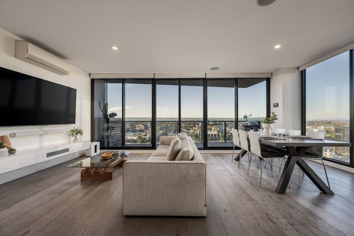 Ein modernes Wohnzimmer mit großem Flachbildfernseher, einer Couch, einem Esstisch mit Stühlen und einem Balkon mit Blick auf die Stadt.