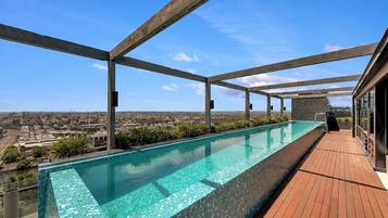 A rooftop infinity pool with a city view, wooden deck, and metal beams.