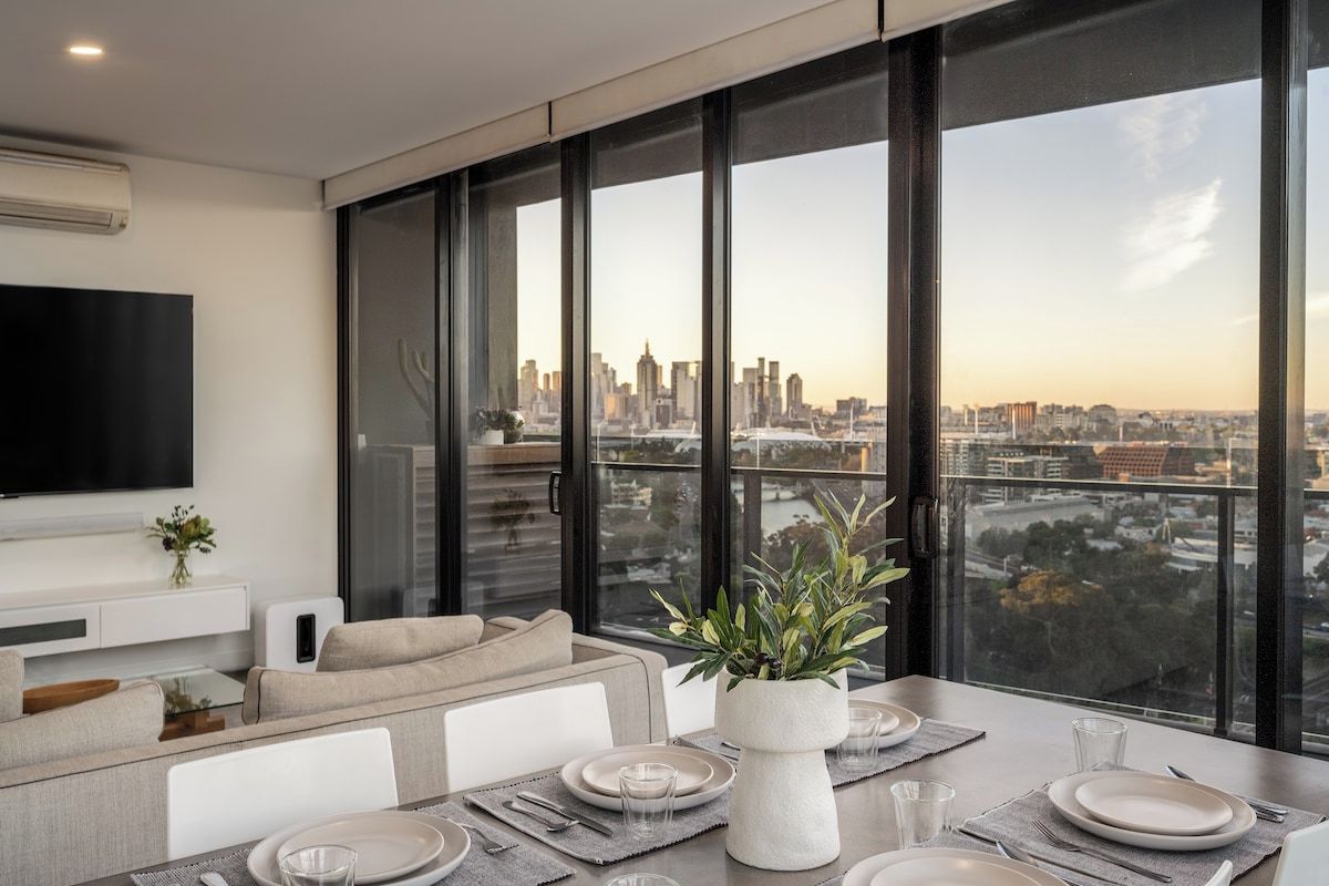 A modern living room with a dining area, large windows, a flat-screen TV, and a city view.