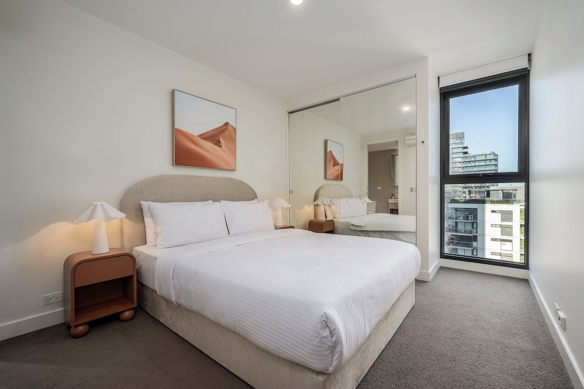 A hotel room with a large bed, a nightstand, a mirror, and a view of the city through a window.