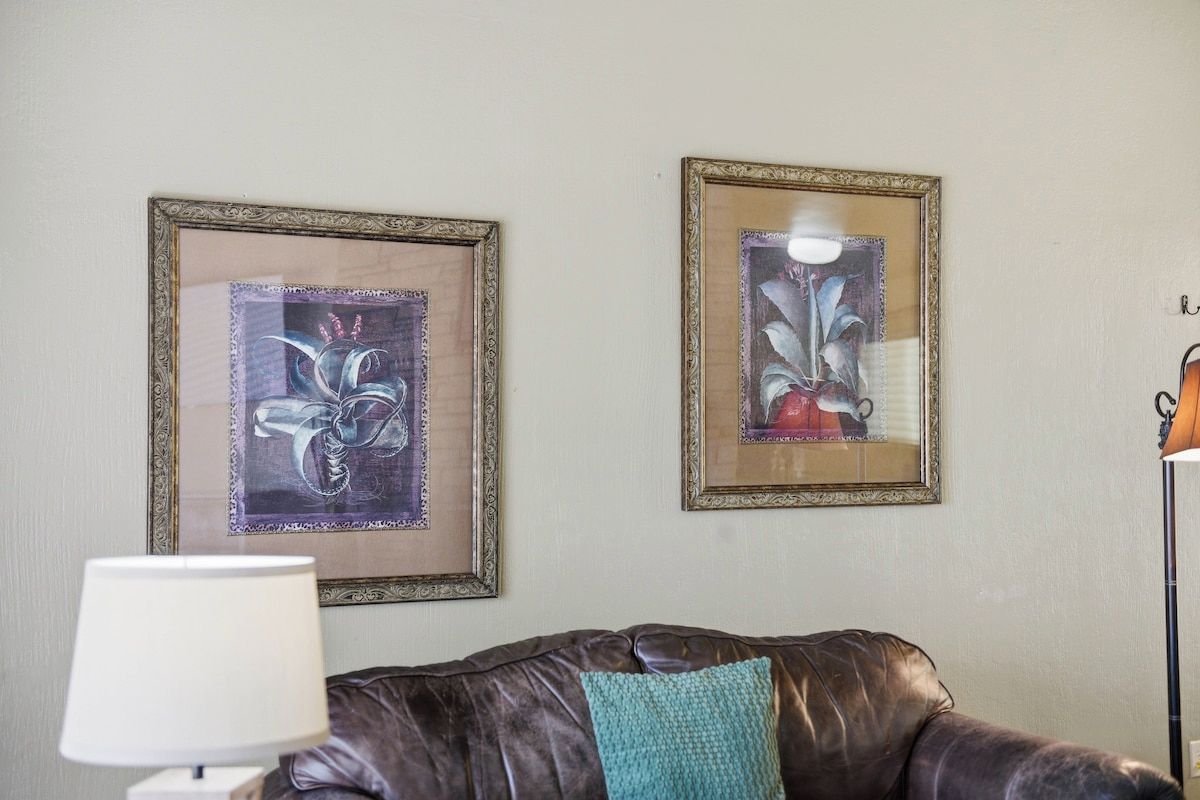 A room with a brown leather sofa, a lamp, and two framed artworks on the wall.