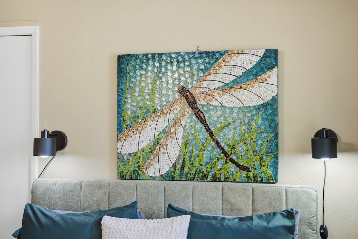 A room with a bed, two wall-mounted lamps, and a mosaic artwork of a dragonfly.