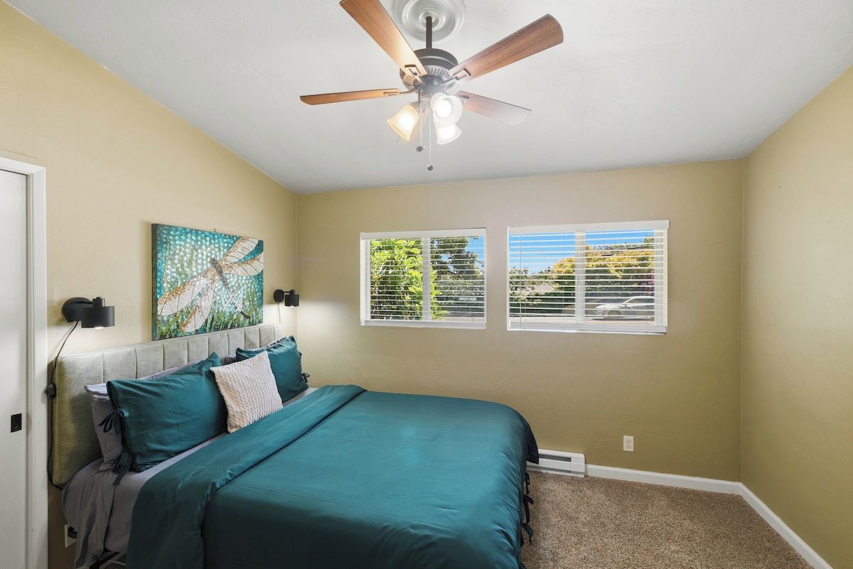 A bedroom with a bed, a ceiling fan, a window with blinds, and a wall-mounted artwork.