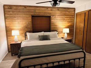 Un dormitorio con panel de madera en la pared, cama con ropa de cama blanca, dos lámparas de noche, ventilador de techo y un armario de madera.