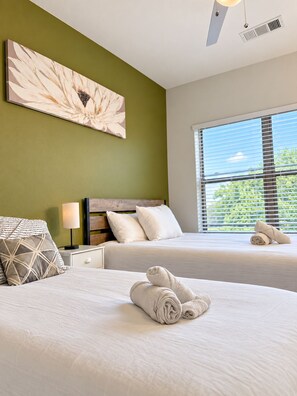 Ein Hotelzimmer mit zwei Betten, einem großen Fenster mit Jalousien, einem Deckenventilator und einem Wandbild.