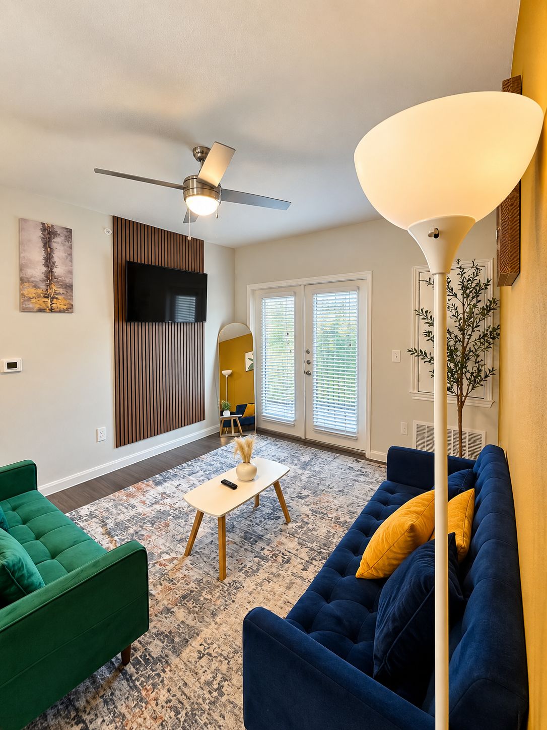 Ein Wohnzimmer mit einem grünen Sofa, einem blauen Sofa, einem Couchtisch, einer Stehlampe, einem Deckenventilator und einem an der Wand befestigten Fernseher.