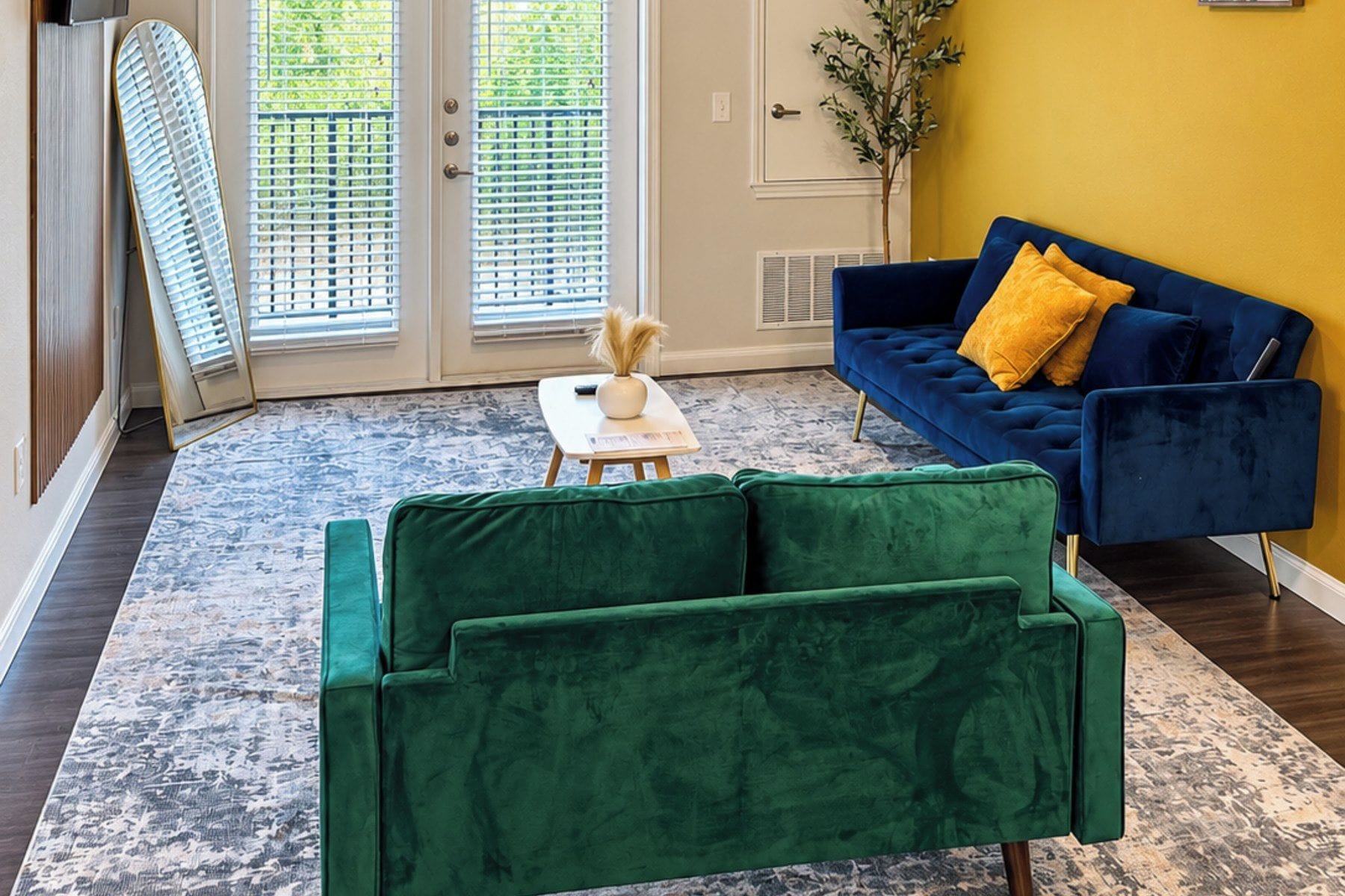 Ein Zimmer mit einem grünen Sofa, einem blauen Sofa mit gelben Kissen, einem kleinen Tisch mit einer Vase und einem gemusterten Teppich.