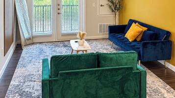 Ein Zimmer mit einem grünen Sofa, einem blauen Sofa mit gelben Kissen, einem kleinen Tisch mit einer Vase und einem gemusterten Teppich.