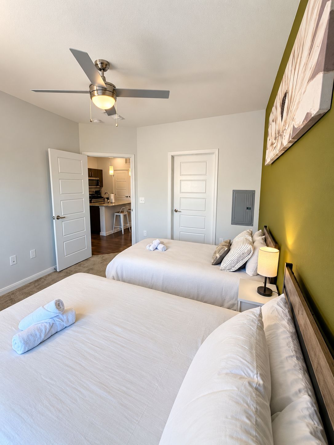 A hotel room with two beds, a ceiling fan, a painting on the wall, and a view of the kitchen through an open door.