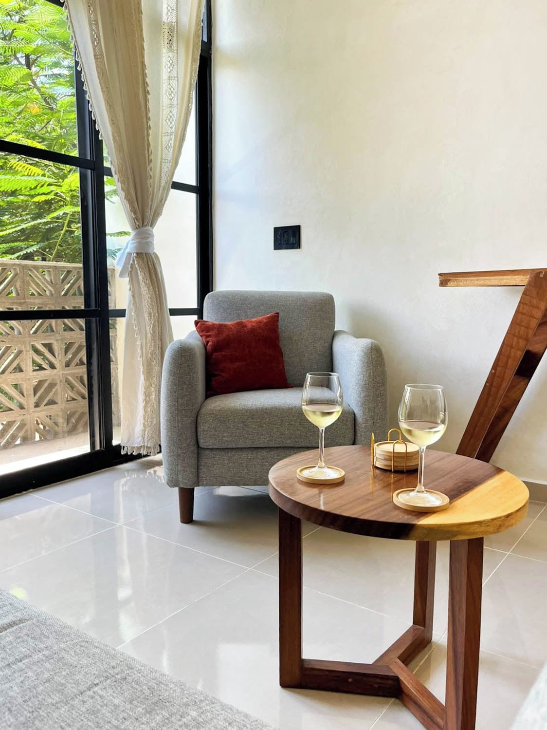 A modern living room with a grey armchair, a small round wooden table with two wine glasses, and a view of greenery through large windows.