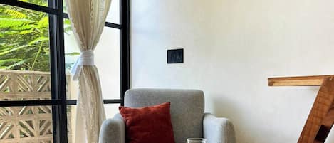 Una sala de estar moderna con un sillón gris, una pequeña mesa redonda de madera con dos copas de vino y vistas a zonas verdes a través de grandes ventanales.