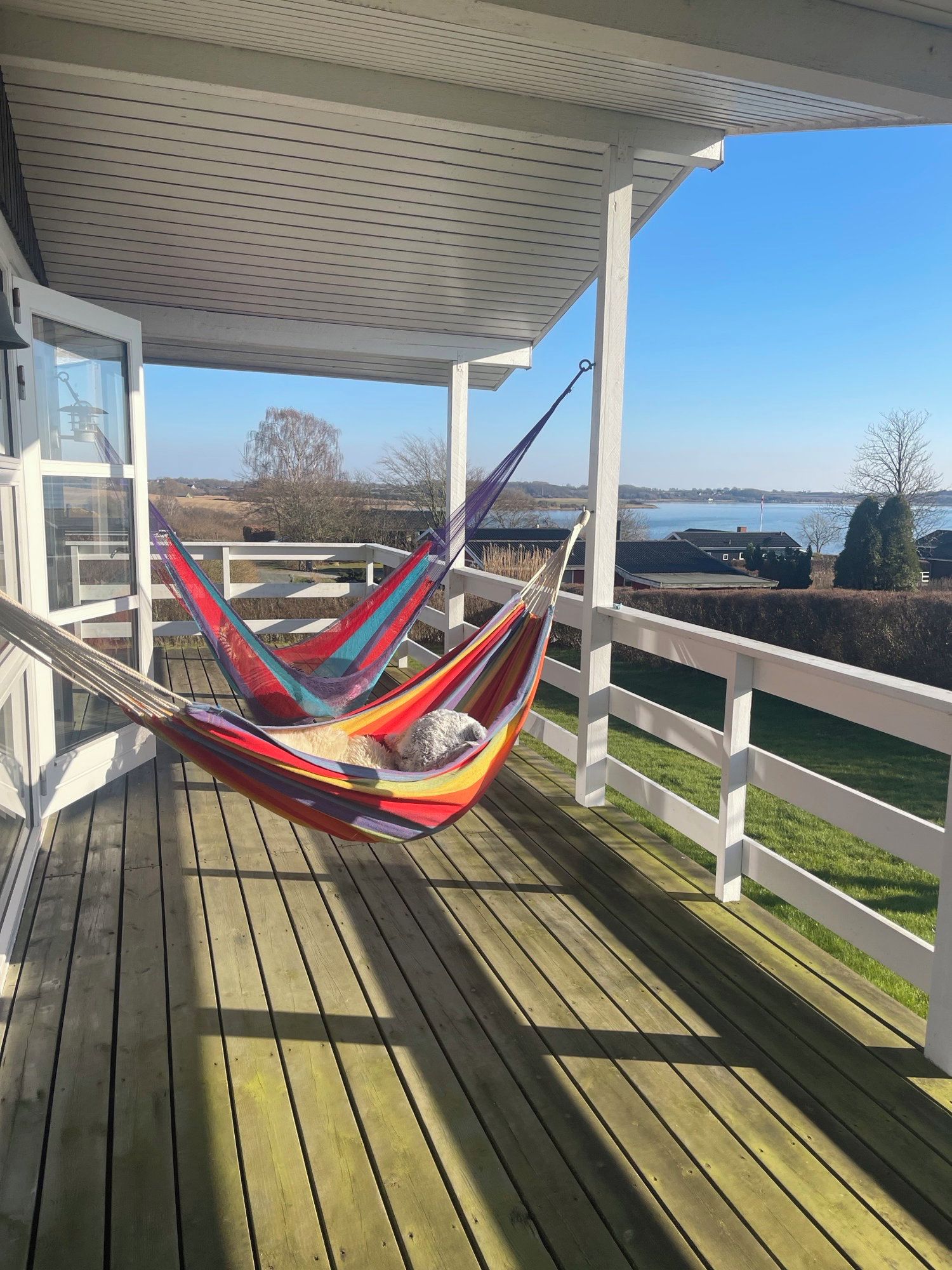 Un hamac sur une terrasse en bois, avec vue sur une étendue d’eau et des bâtiments éloignés.