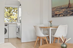 A modern kitchen with a washing machine, a dining area with a table and chairs, and a cityscape painting on the wall. - The Primrose Loft | Garden Views | Regents Park (Greater London)