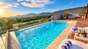 A rooftop infinity pool with clear blue water, surrounded by a glass railing, and overlooking a mountainous landscape.
