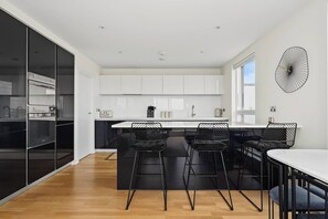 A modern kitchen with white cabinets, a central island with bar stools, and a decorative wall clock. - The Masson Terrace | 3-Bed | Parking & Kew Gardens (Greater London)