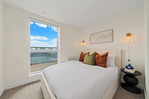Et hotelværelse med en stor seng, et vindue med udsigt til bygninger, et natbord med en lampe og et billede i ramme på væggen.