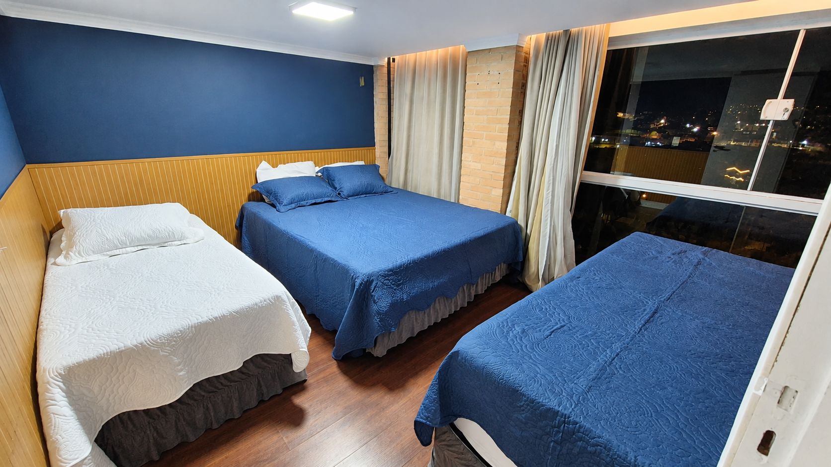 Une chambre d’hôtel avec trois lits, une grande fenêtre avec des rideaux et une vue sur la ville.