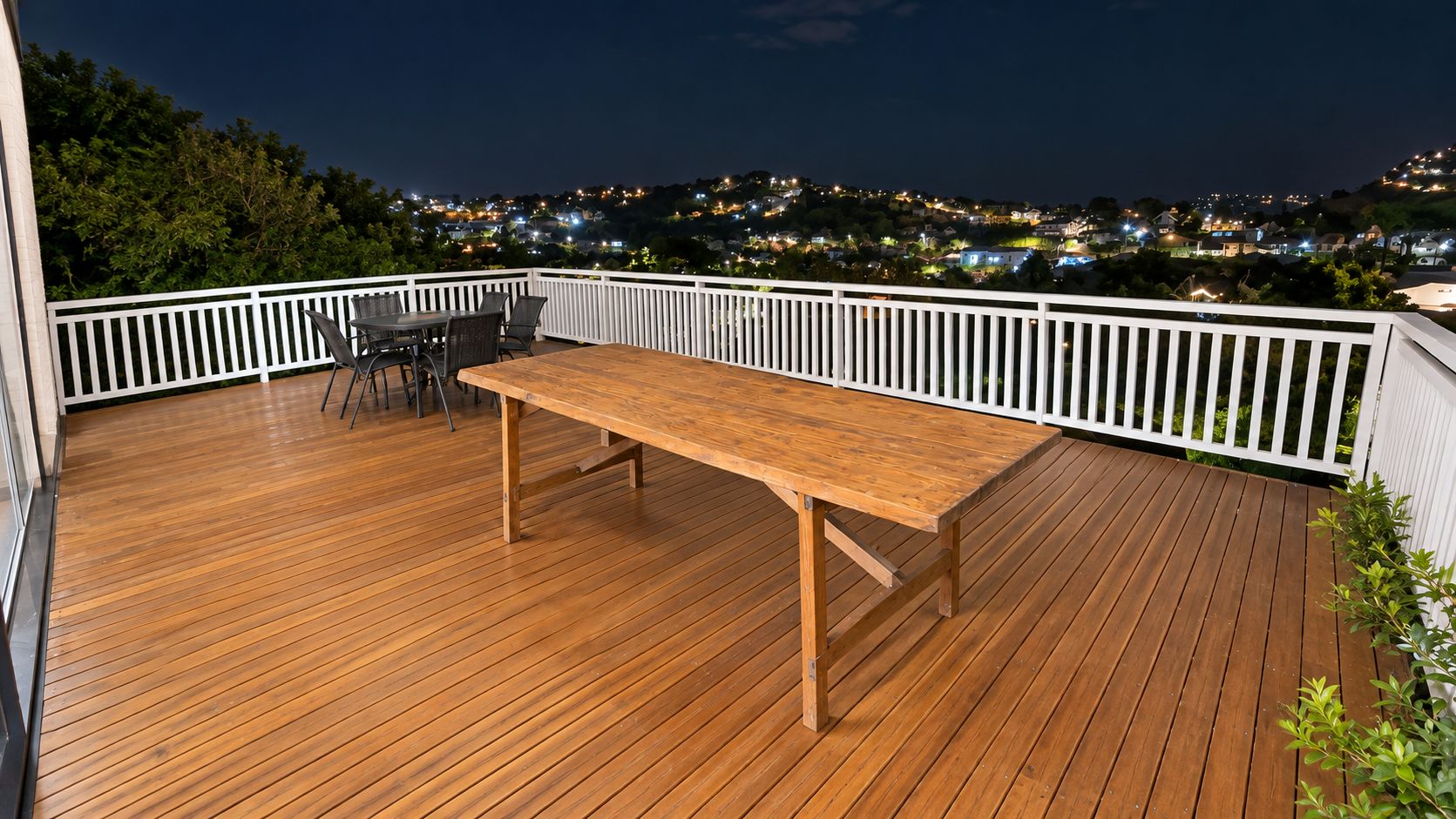 Eine Holzterrasse mit Tisch und Stühlen, umgeben von einem weißen Geländer, mit Blick auf eine nächtliche Skyline.