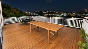 En träveranda med ett bord och stolar, omgiven av ett vitt räcke, med utsikt över en stadssiluett i nattens mörker.