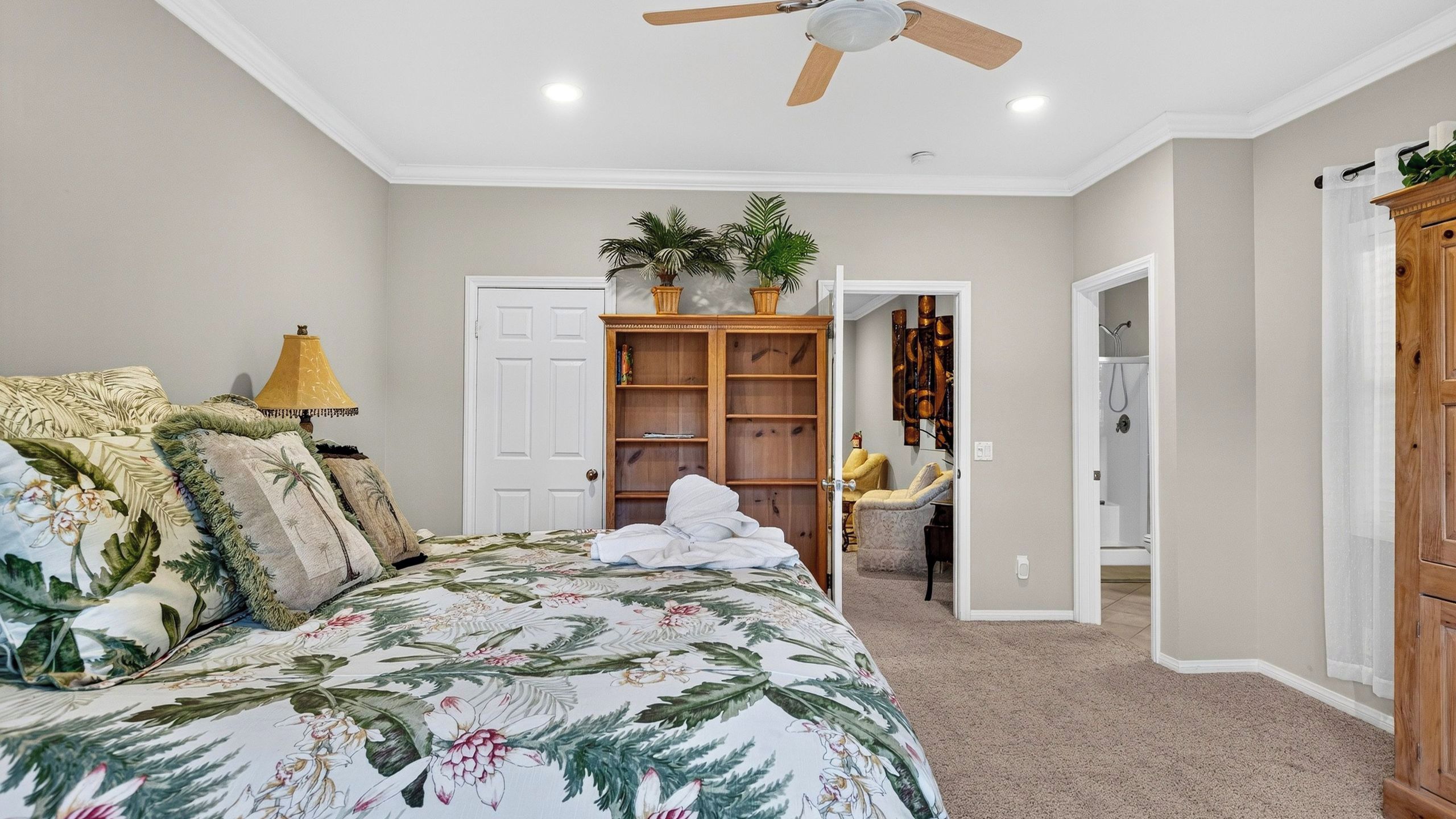 A bedroom with a bed, a ceiling fan, a wooden wardrobe, a bathroom with a shower, and a hallway leading to another room.