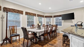 A dining area with a table set for six, flanked by patterned chairs, with large windows and a flat-screen TV mounted on the wall. - Wooded Wonder by Avantstay Game Room + Covered Decks + Fire Pit Near Lake Arrowhead (Lake Arrowhead)