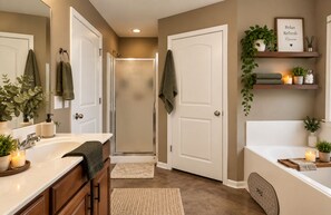 Une salle de bain avec une douche, une baignoire, un meuble-lavabo avec un miroir et une étagère décorée de plantes et de bougies.
