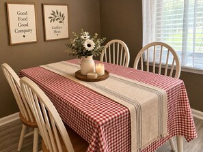 Un coin repas avec une nappe à carreaux, des chaises blanches, un vase de fleurs et des panneaux encadrés accrochés au mur.
