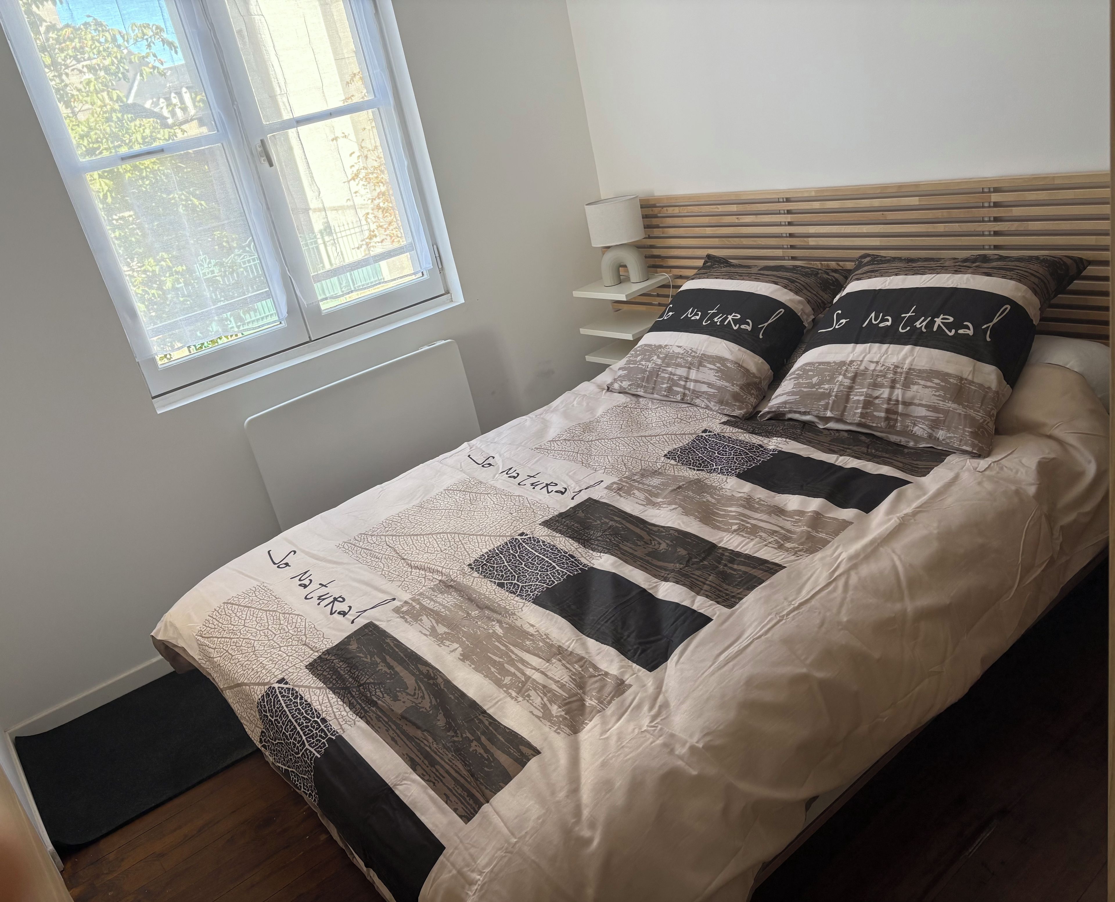 Un letto con testiera trapuntata, un comodino con lampada e una finestra vista alberi.