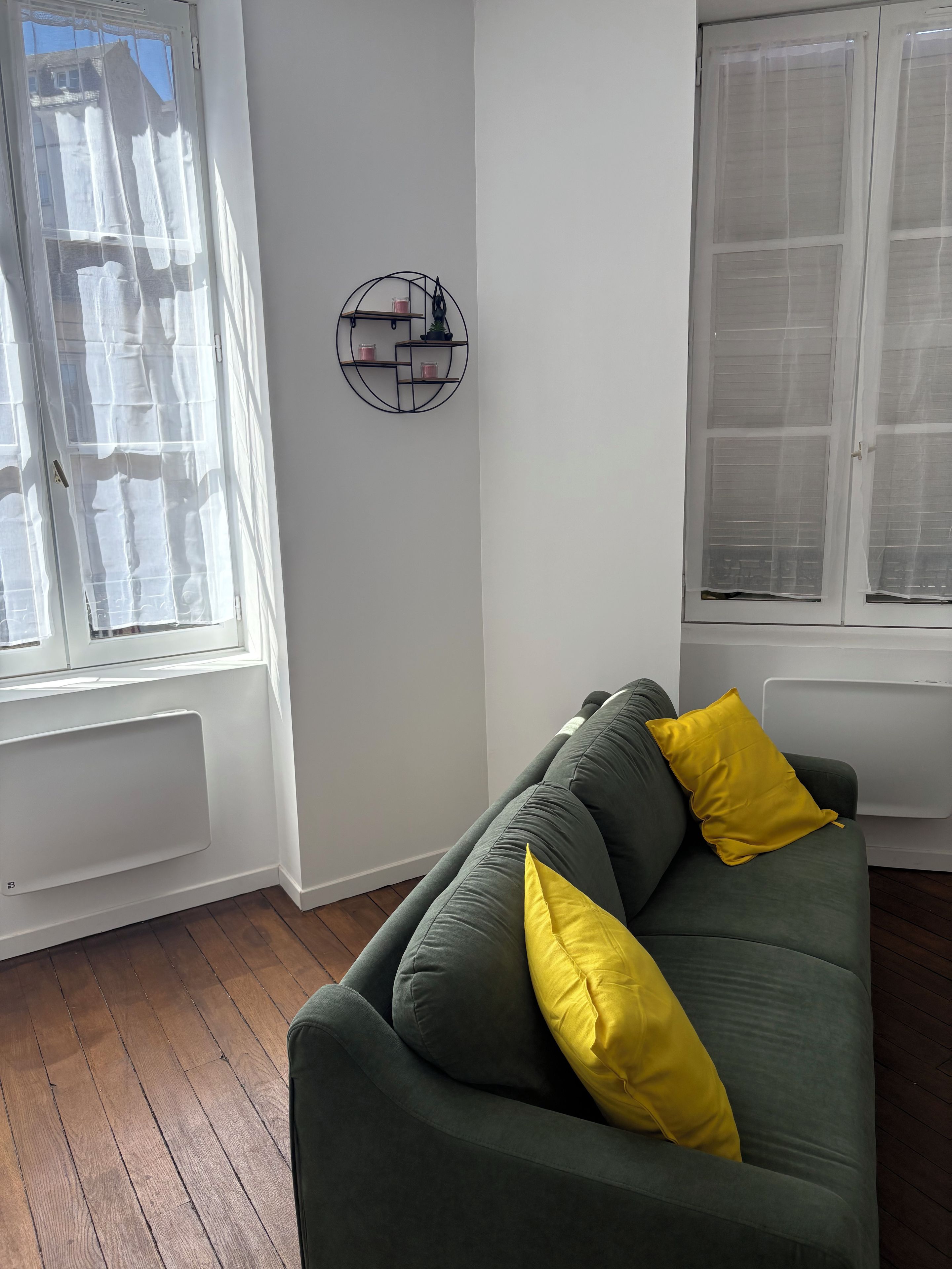 A room with a green sofa, a yellow pillow, a wall-mounted shelf with items, and a window with curtains.
