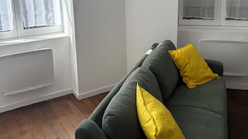 A room with a green sofa, a yellow pillow, a wall-mounted shelf with items, and a window with curtains.