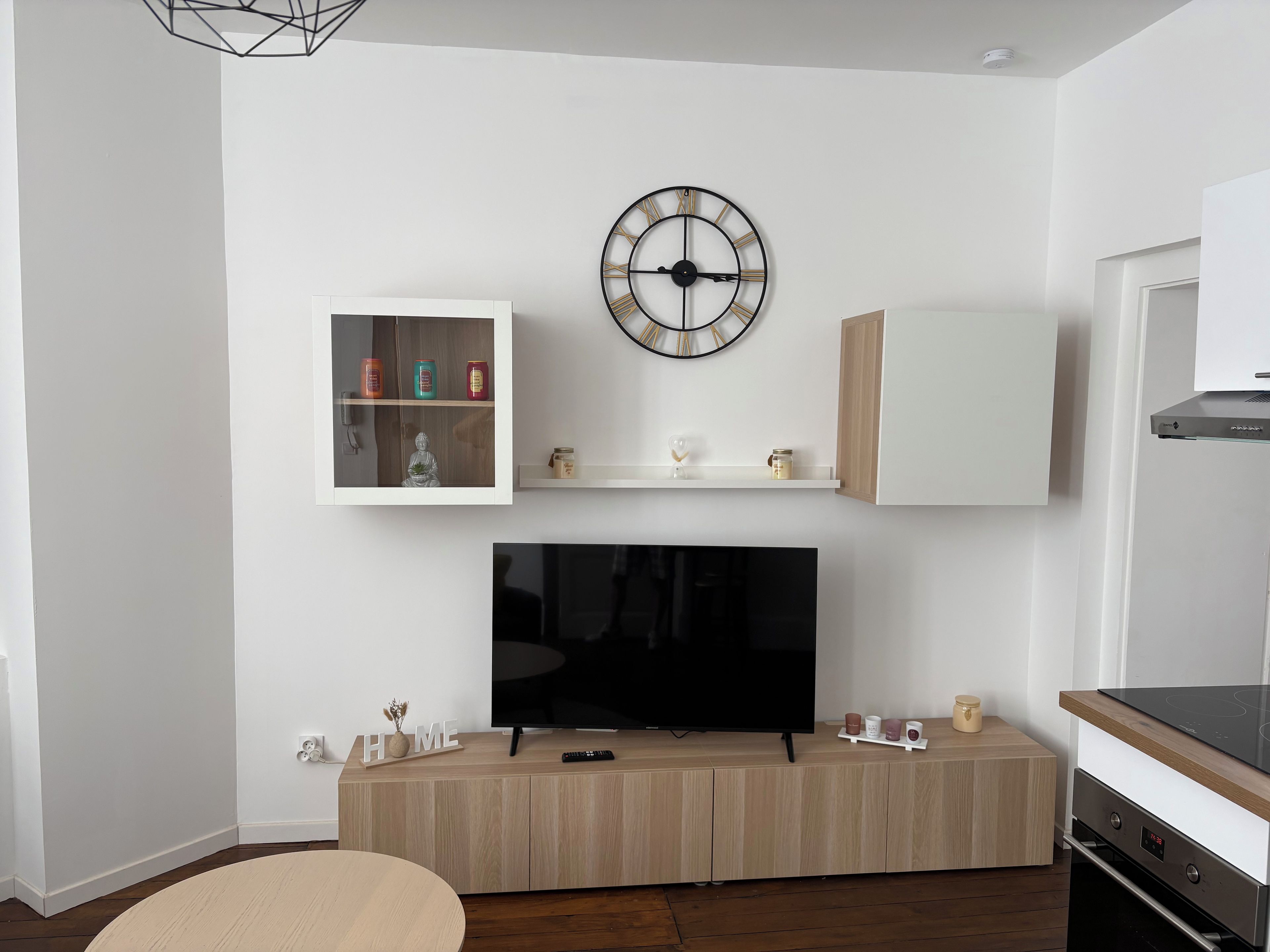 Een moderne woonkamer met een flatscreen televisie, houten kasten, een hangend horloge en een klein plankje met decoratieve objecten.