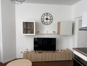 A modern living room with a flat-screen TV, wooden cabinets, a mounted clock, and a small shelf with decorative items. - New Listing (Limoges)