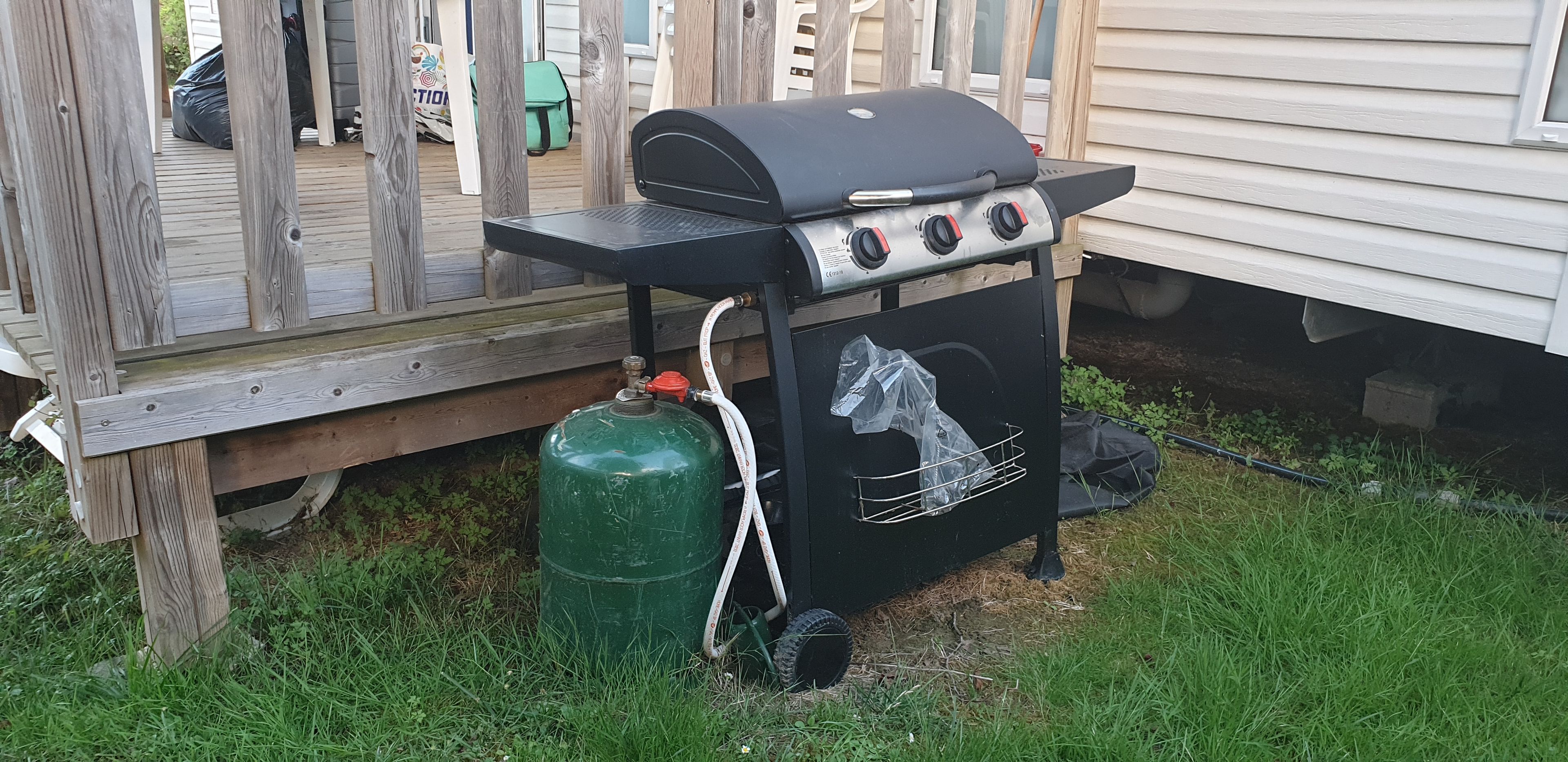 Una parrilla para barbacoa con un tanque de gas y una manguera, ubicada sobre una terraza de madera.
