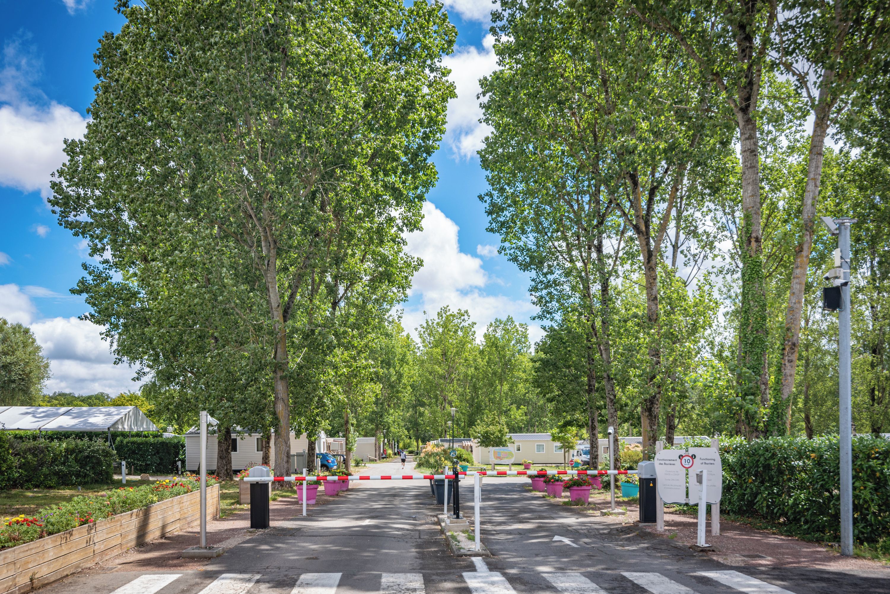 A gated entrance with a pedestrian crossing, surrounded by trees and a clear sky.