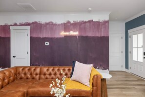Ein Zimmer mit einem braunen Ledersofa, einer Wand mit einem abstrakten Design in Lila und Rosa, und einem Neonlicht mit der Aufschrift 'Aorn Street'.