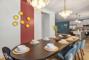 Ruokailutilassa on pitkä pöytä, johon on katettu usealle henkilölle, taustalla näkyy keittiö ja seinää koristavat pyöreät esineet.