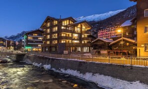 Un hotel de varios pisos con balcones, un río al frente y montañas nevadas al fondo.