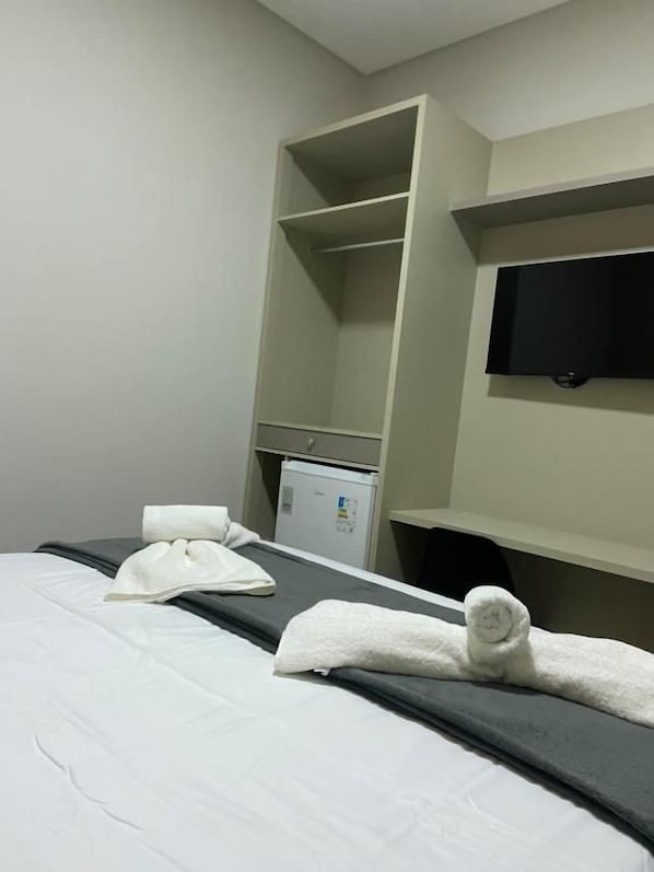 A hotel room with a bed, a flat-screen TV mounted on the wall, a minibar, and a built-in shelving unit.