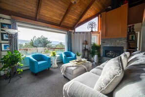 Una sala de estar acogedora con chimenea, un sillón azul, un sofá blanco y vistas al exterior.