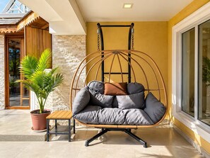 A hanging chair with cushions, a potted plant, and a wooden table in a sunlit outdoor area.