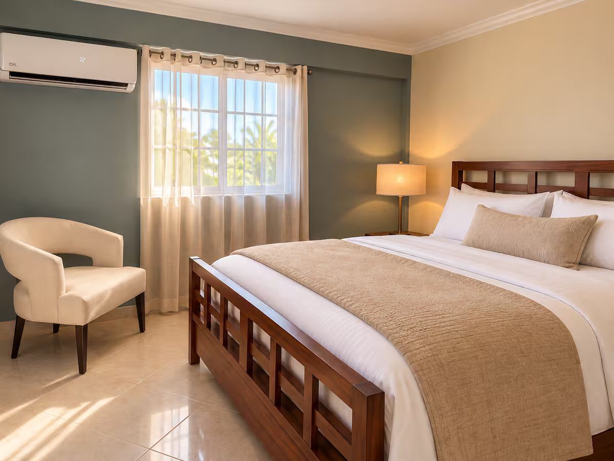 A hotel room with a bed, a chair, a lamp, and an air conditioning unit.