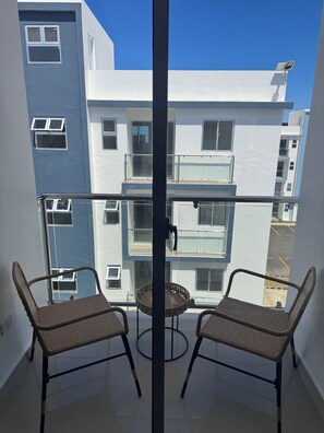 A balcony with two wicker chairs, a glass table, and a view of a multi-story building with balconies. - Brand-New Modern Retreat in Sosúa (Sosua)