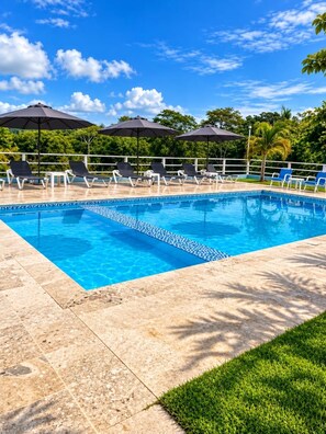 A swimming pool with lounge chairs and umbrellas, surrounded by a tiled deck and greenery. - Brand-New Modern Retreat in Sosúa (Sosua)