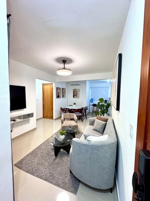 A modern living room with a flat-screen TV, a grey sofa, a coffee table, and a dining area in the background. - Brand-New Modern Retreat in Sosúa (Sosua)
