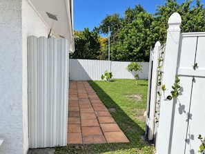 Un camí pavimentat que condueix a una tanca blanca, amb un arbre petit i plantes en testos.