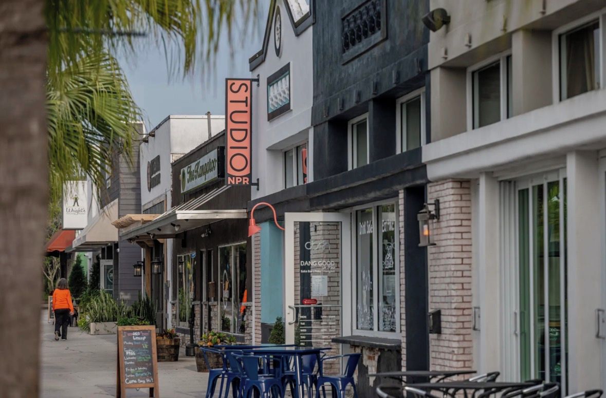 Vista de la calle con un edificio identificado como 'STUDIO NPR' y otro con la leyenda 'DANG GOOD'. Se observan zonas de estar al aire libre con mesas y sillas.