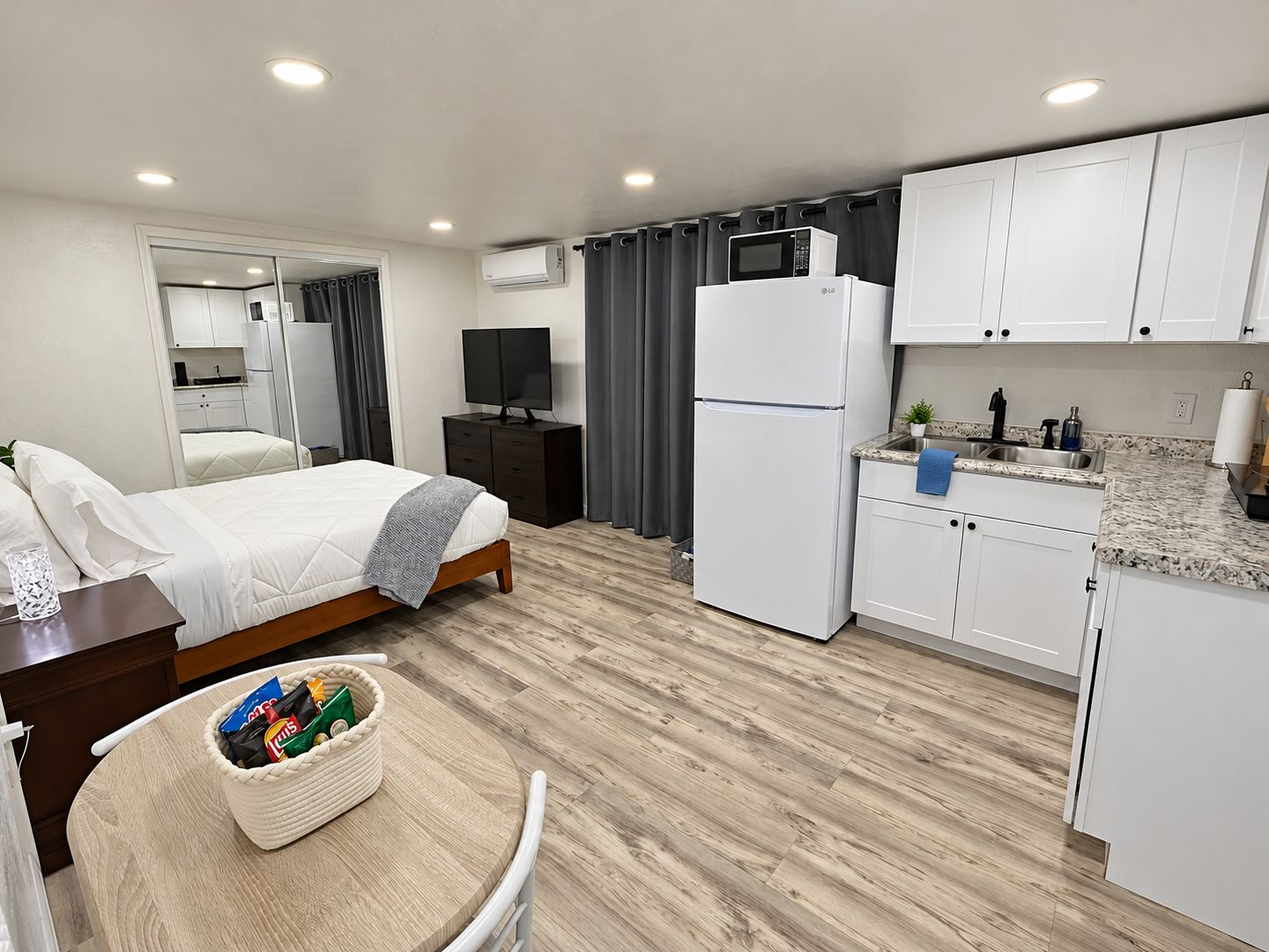 A compact hotel room with a bed, bedside table, TV, kitchenette, and a small dining area.