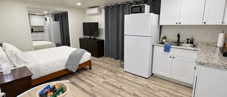 A compact hotel room with a bed, bedside table, TV, kitchenette, and a small dining area.